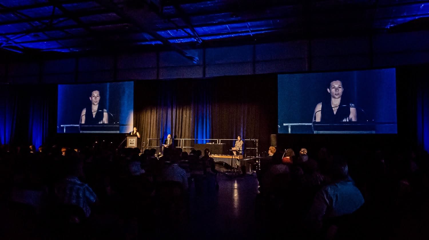 Kelly delivering the Helena College graduation keynote on stage, shown on big screens